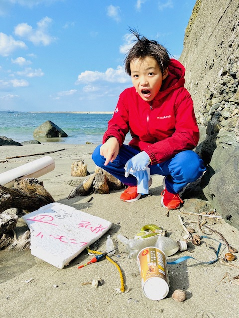 青空と青い海の見える砂浜。ポイ捨てされたのか、たくさんのごみを指さしている男児の写真。