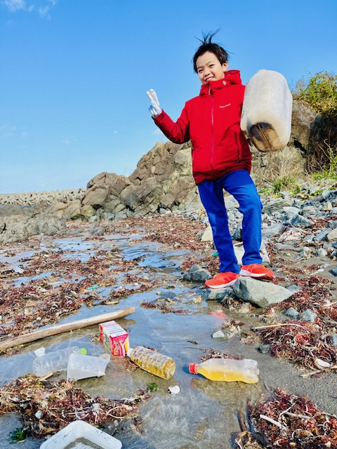 海藻やペットボトルの漂着ごみが多い岩場。男児がプラスチックごみを手に持っている写真。
