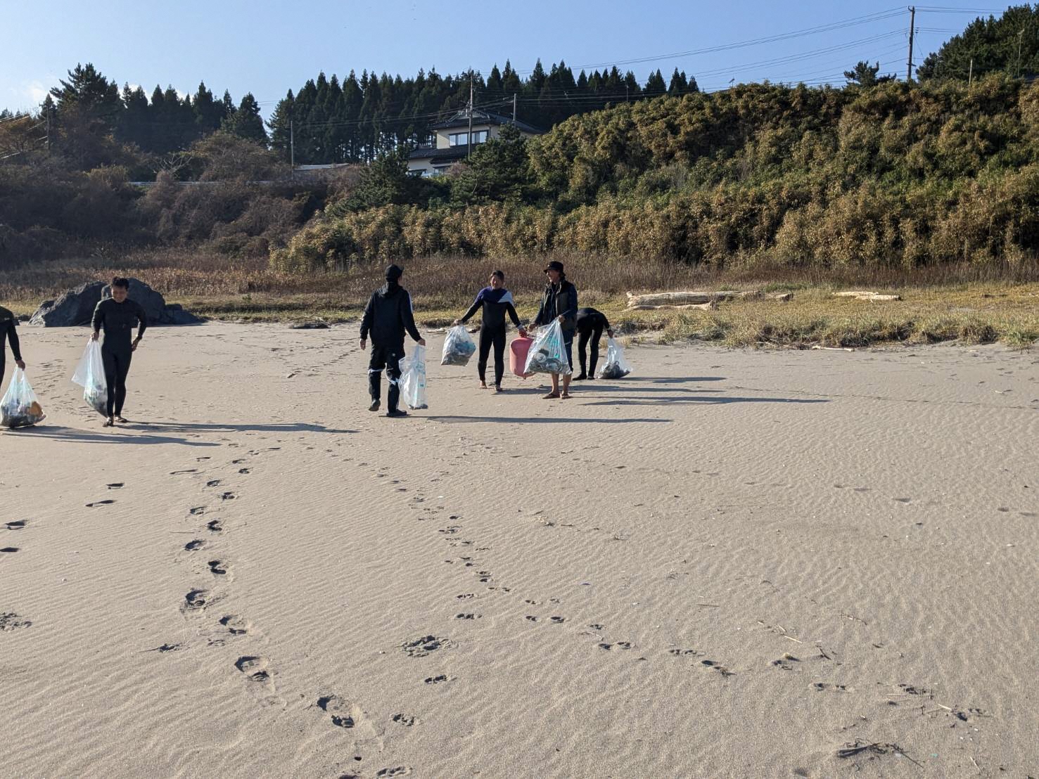 奥に家と木々の見える手前の砂浜に、足あとが続いている。参加者がごみの入っている袋を手に持っている。