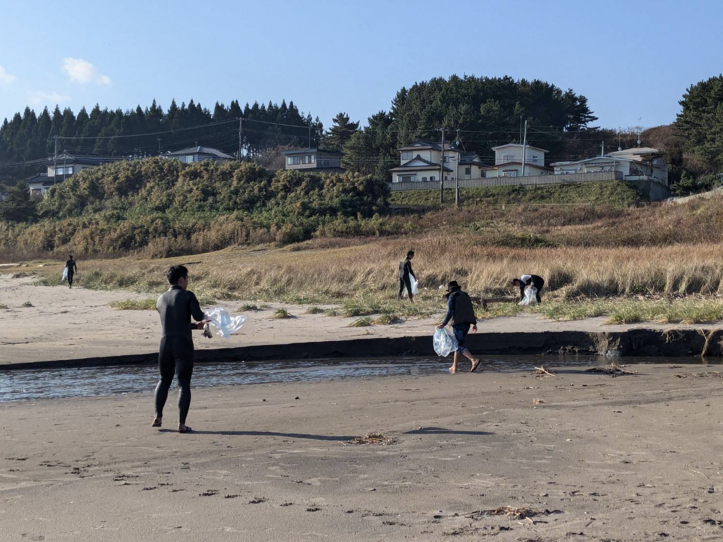 奥に住宅や森が広がっている。砂浜の間に海の流れている場所で集めたごみを袋に入れている参加者の写真。