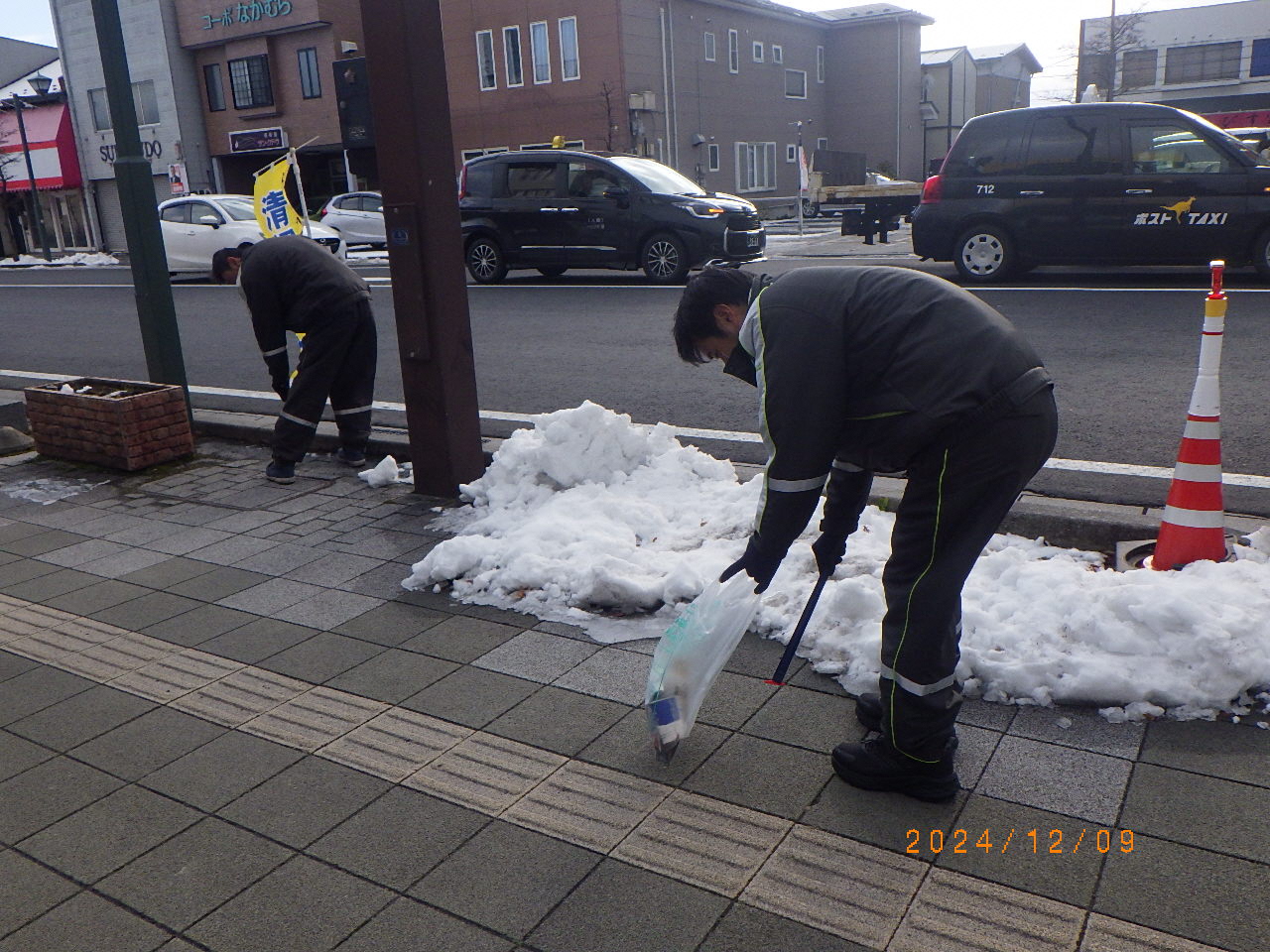 雪のある歩道のごみをトングで拾っている男性二人の写真。