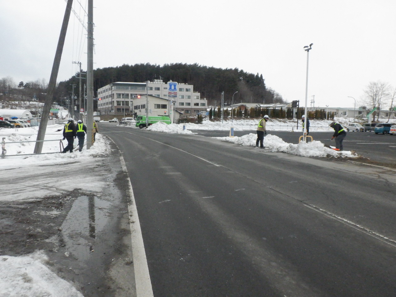 遠くにコンビニエンスストアの看板の見える道路。歩道で防寒服、ヘルメット、反射ベストを着用し活動する人たち。