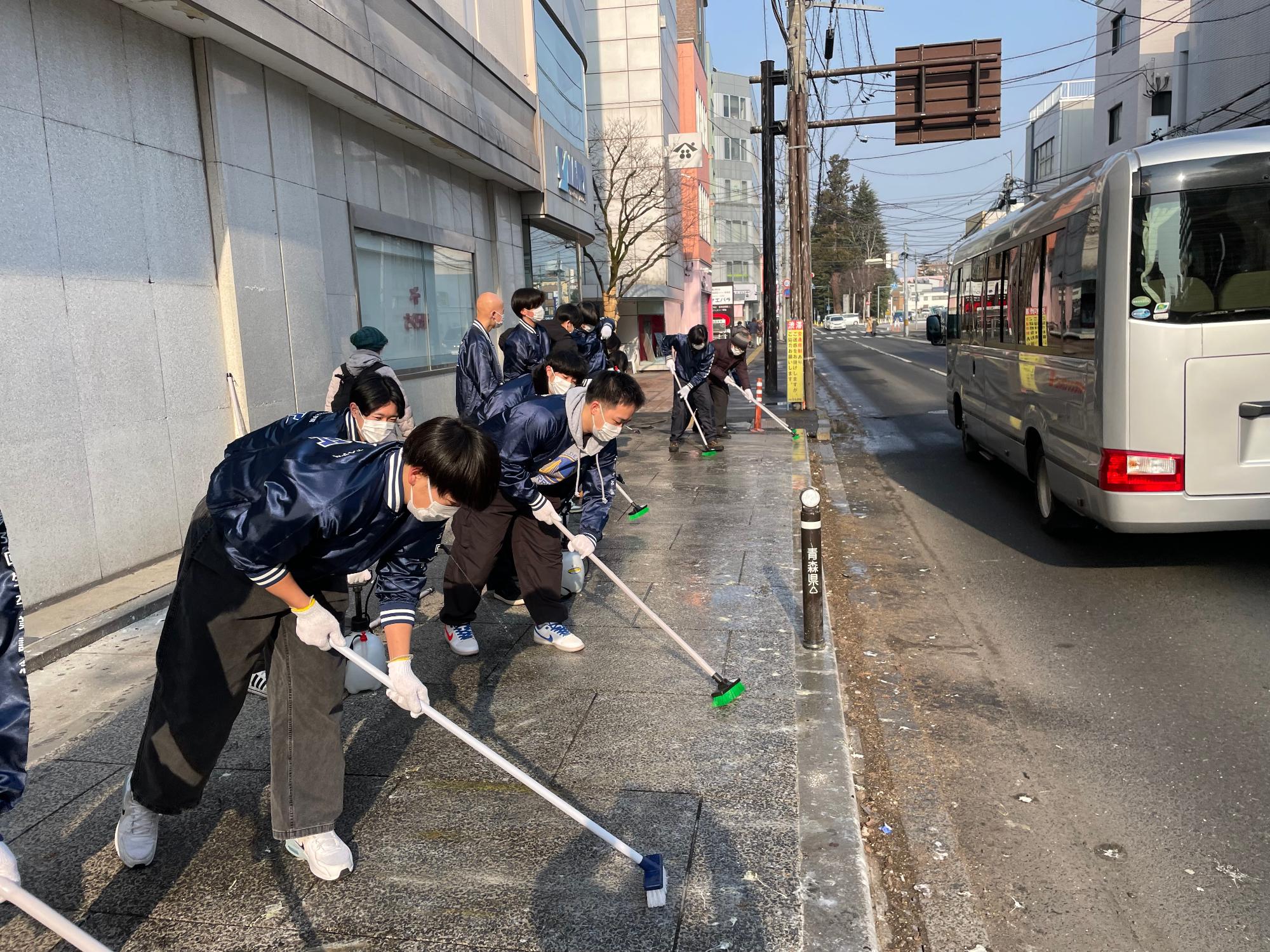 中心街の鳥のフンで汚れた歩道を、デッキブラシで掃除する10名くらいの参加者の写真