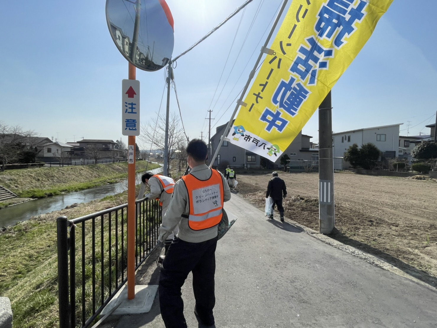 清掃活動中ののぼりを掲げ浅水川沿いの道路のごみを拾っている写真