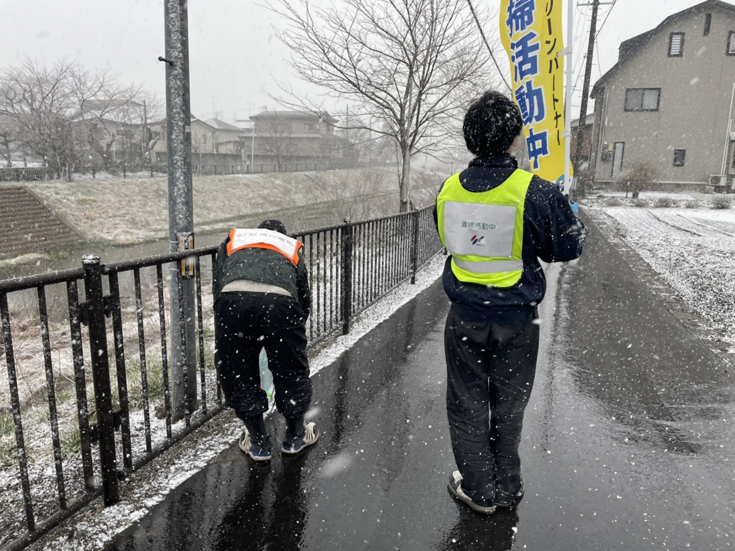 雪が舞う中のぼりをかかげながら、道路のごみを拾う男性二人の写真