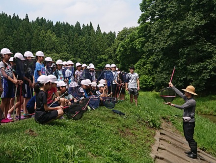 小川での活動前に、講師が川岸で生き物の取り方や注意事項を説明し、こども達が話を聞いている様子の写真