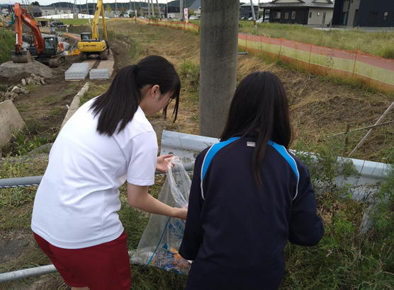 ショベルカーの見える場所でボランティア用ごみ袋に拾ったごみを入れている女子生徒二人の写真