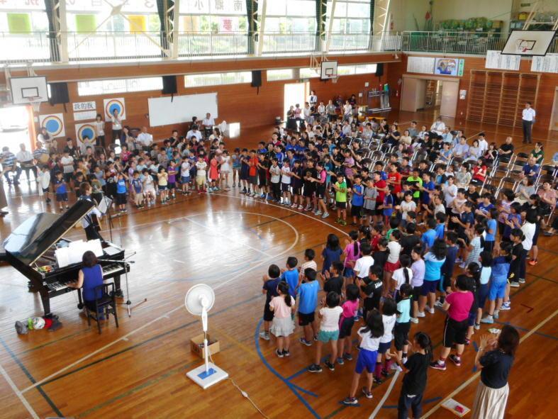 ピアノを弾いている豊嶋さんと少し離れた位置に立っている手拍子をしている児童たちを上方から撮影した体育館内の写真