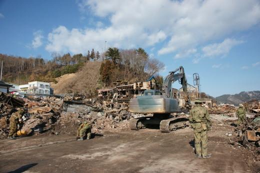平成23年3月18日：左に瓦礫の山、中央の道路でショベルカーが瓦礫を撤去している、手前に自衛隊員が立っている写真