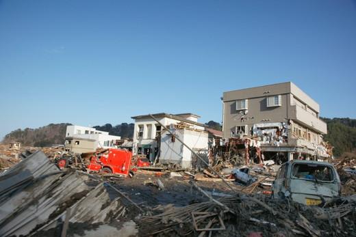 平成23年3月18日：手前に瓦礫や軽自動車、赤い車が写っている、奥に住宅が3棟立っている写真