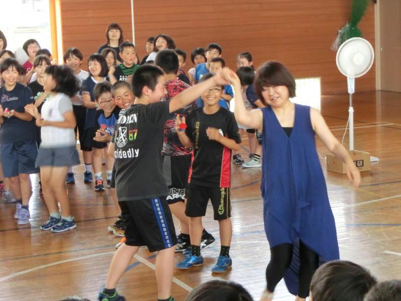 奥に笑顔の児童たちと、手前で手を取り合ってダンスをしている男子児童と豊嶋さんの写真