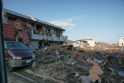 平成23年3月18日：左手前に茶色のバンタイプの自動車、左奥にアパート、中央には瓦礫の山が写っている写真
