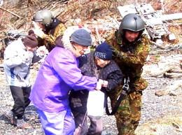 瓦礫が散乱するなか、中央の年配女性を介抱しながら歩く左に紫色の服をきた男性、右に自衛隊員の写真