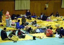 体育館の床にしかれた毛布の上に寝転んだり座ったりしている宿泊者たちの写真