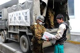 災害派遣の垂れ幕のかかった自衛隊車両の前で自衛隊員から物資を受け取るらう男の子の写真