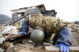 瓦礫をのぞき込む自衛隊員、奥には被災した住宅が立っている写真