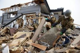 瓦礫が散乱しているなか瓦礫を撤去する自衛隊員、奥には被災した住宅の写真
