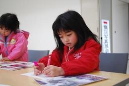 左手で鉛筆をもって座って手紙を書いている赤い服を着た女の子の写真