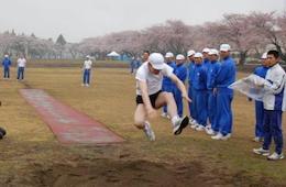 体操服を着て走り幅跳びのジャンプしている人、右側に青ジャージを着て並んでいる人たちの写真