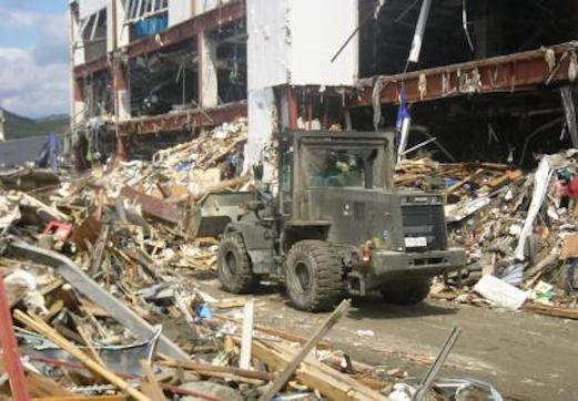 瓦礫が散乱している中をブルドーザーが瓦礫を撤去している、後ろには崩れかけた建物が写っている写真