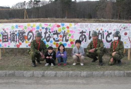 左に1名、右に2名の自衛隊員、真ん中に3名の子供が座っている、後ろには「自衛隊のみなさんありがとう」の垂れ幕が写っての記念写真