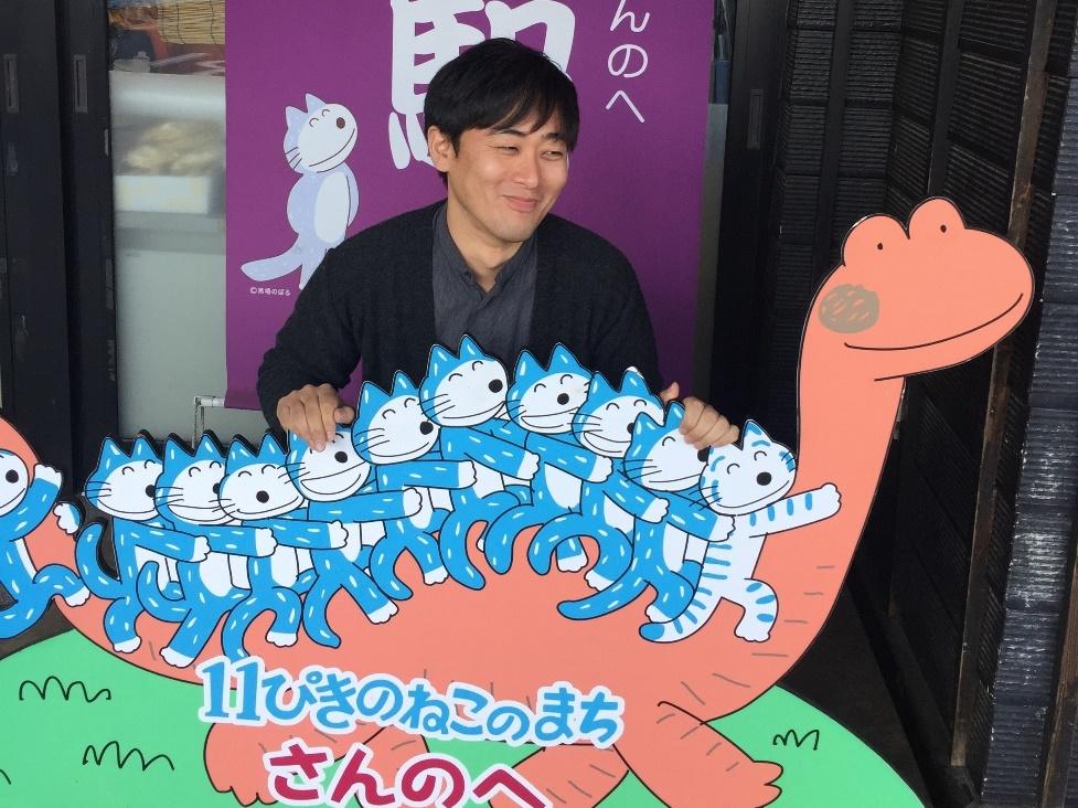 11匹のねこのまち、の11匹の猫と恐竜が描かれたイラストを手に、笑顔の出川 友久さんの写真