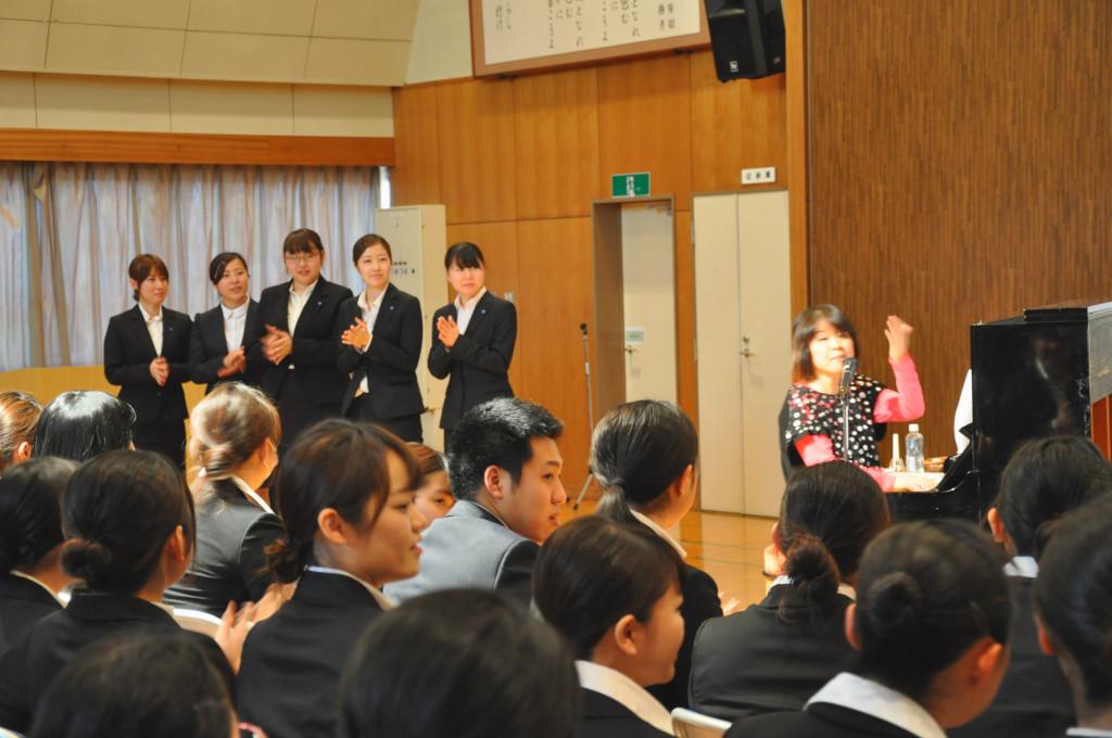 手前に着席している生徒たち、前に立って手拍子をしている5人の生徒たちとピアノを弾いて歌っている豊嶋さんの写真