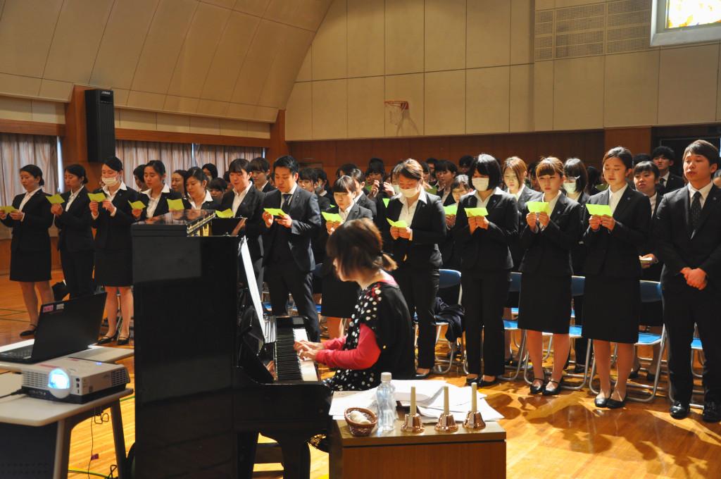 ピアノを弾いている豊嶋さんと、歌詞カードを見ながら立って歌っている生徒たちの写真