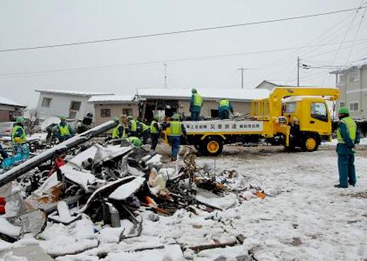 手前に雪が積もっている瓦礫、奥で撤去作業をしている人たちとトラックの写真