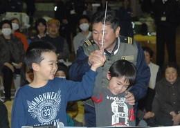 男子児童2名に指揮棒の振り方を教えている自衛隊の作業着を着た男性の写真