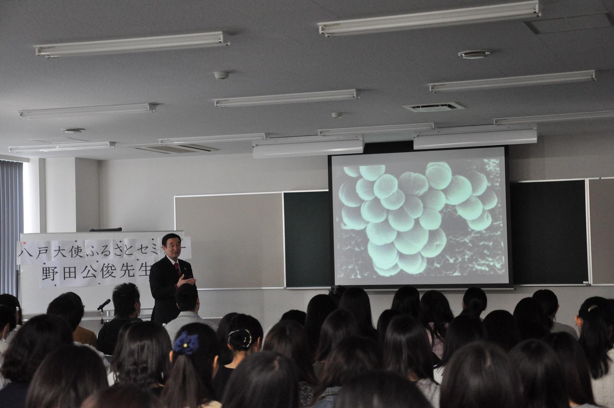 モニターに映された細菌の画像を見ながら話をしている野田先生と、着席して話を聞いている生徒たちの後姿の写真