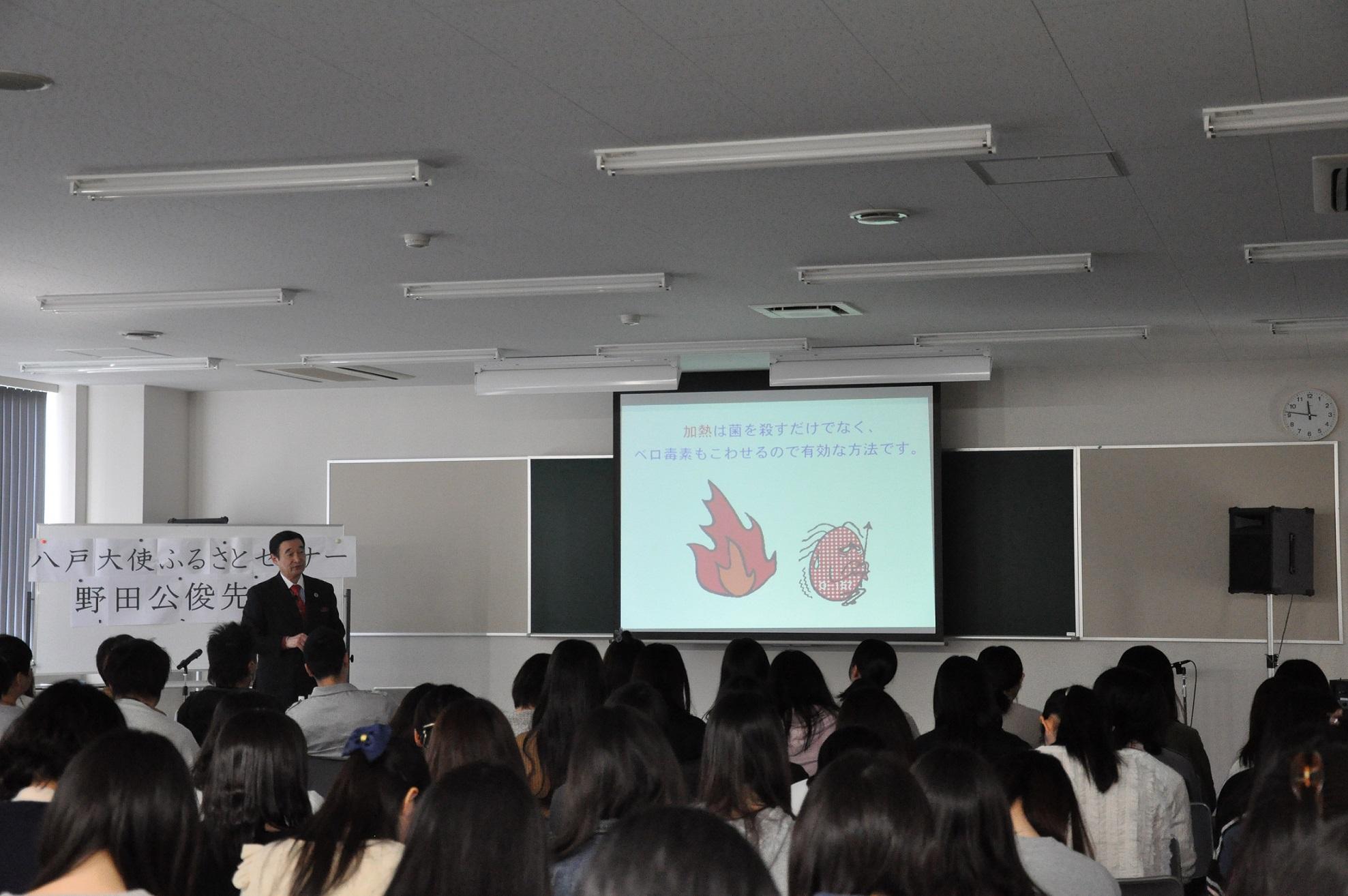 モニターに写っている画像を見ながら話をしている野田先生と、着席して話を聞いている生徒たちの後姿の写真