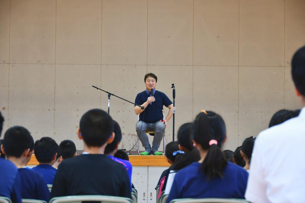 椅子に座って並ぶ生徒たちを前に、ステージ上でマイクを持って自己紹介をする松田さんの写真