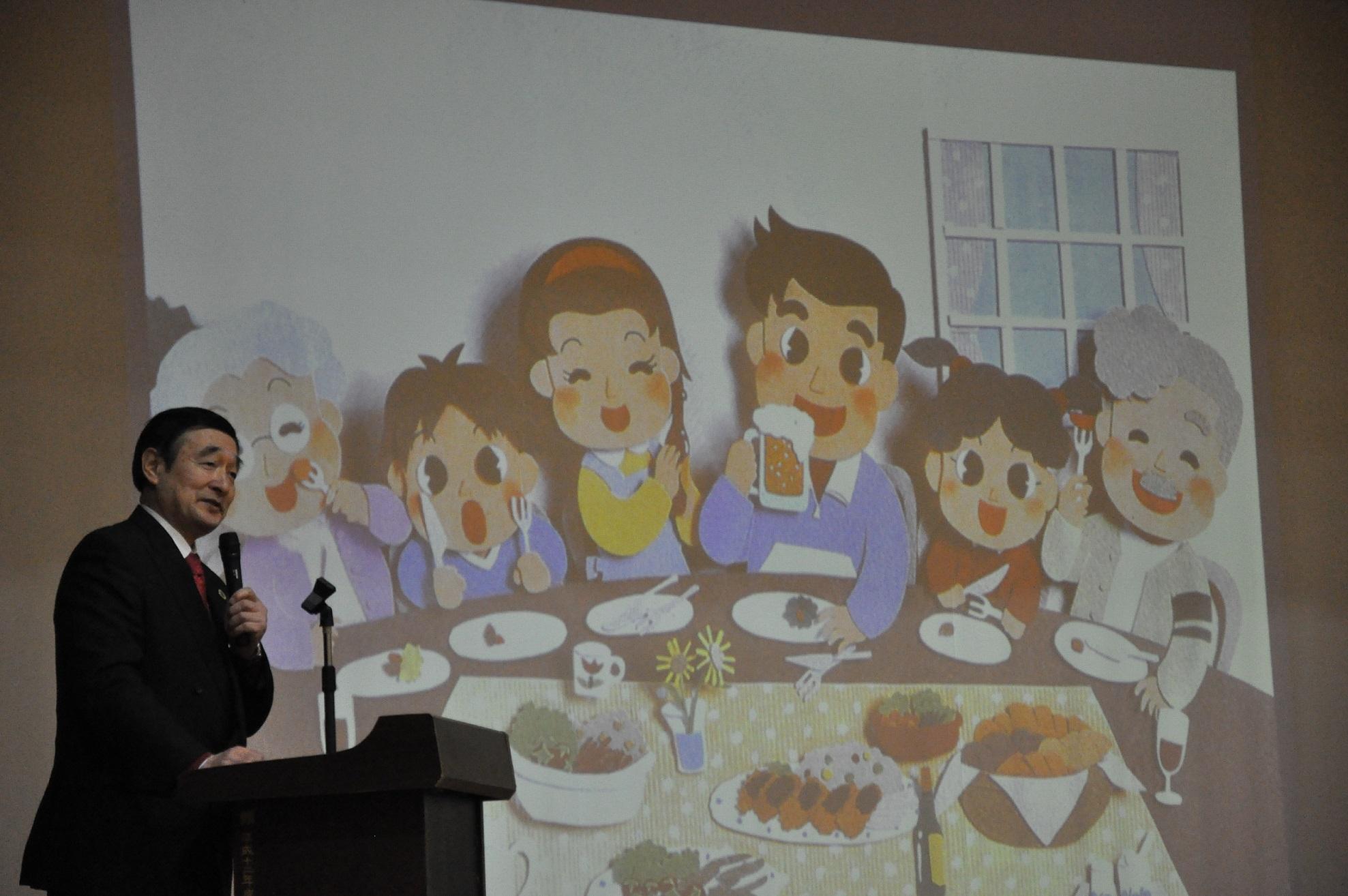 嘱託を囲む家族のイラストの写っているスクリーンの横で、マイクを持って話をしている野田先生の写真