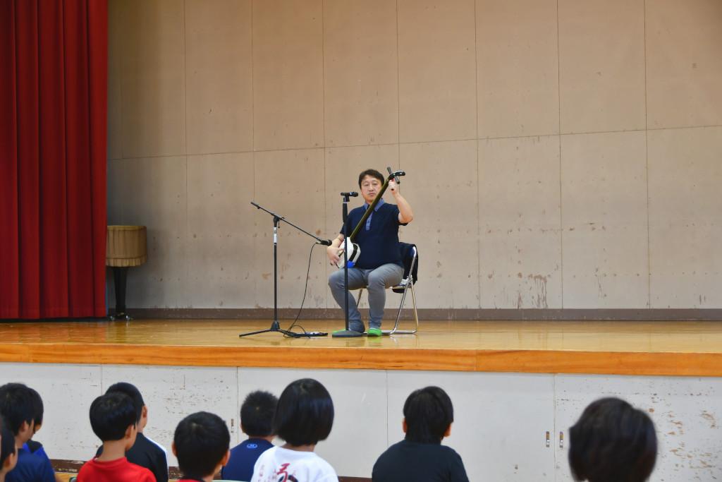 椅子に座って並ぶ生徒たちを前に、ステージ上で三味線の演奏を披露する松田さんの写真
