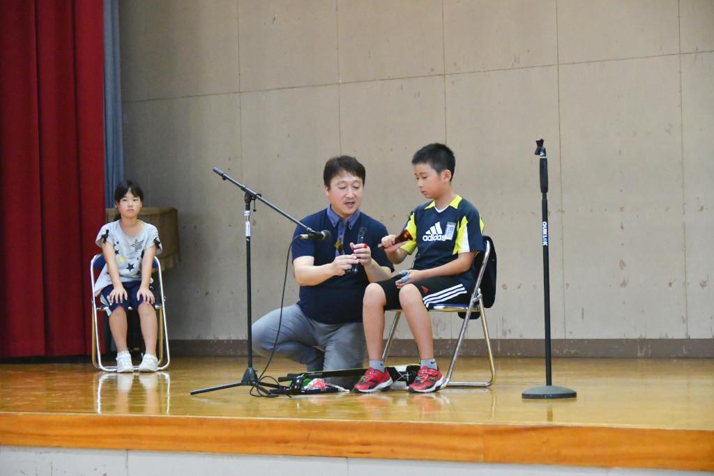 ステージ上で男子生徒に三味線体験のレクチャーを行う松田さんと少し離れた椅子で順番を待つ女子生徒の写真