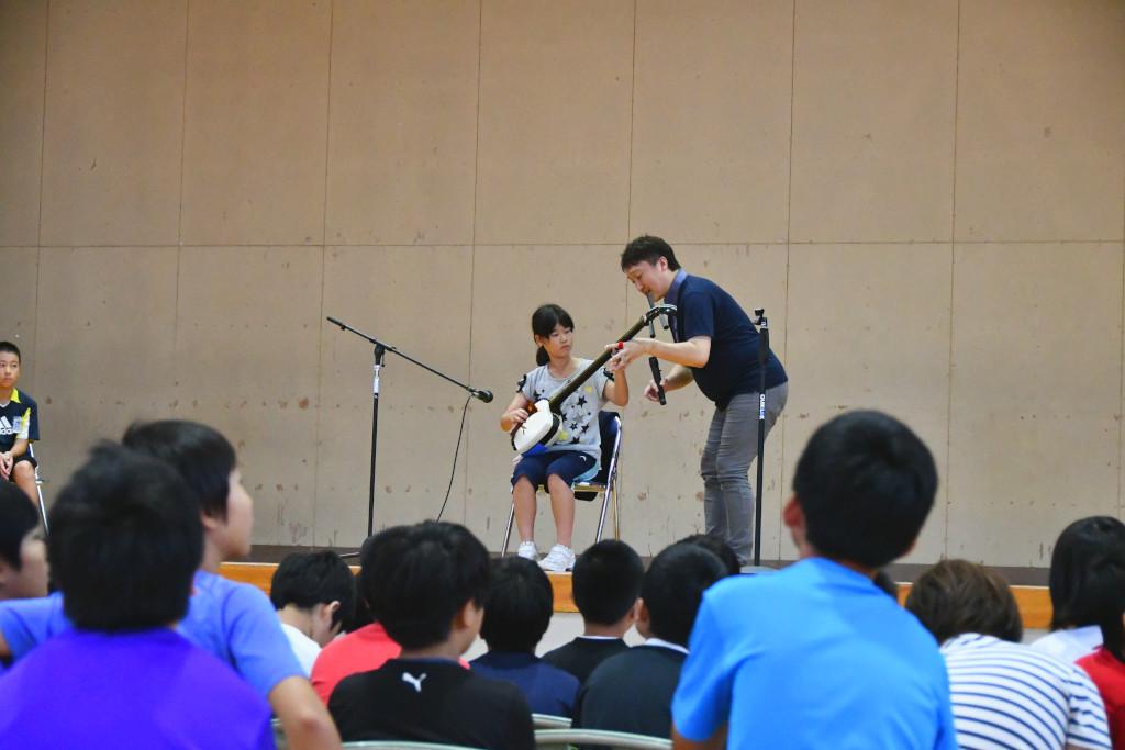 ステージ上で女子生徒に三味線体験のレクャーを行う松田さんの写真