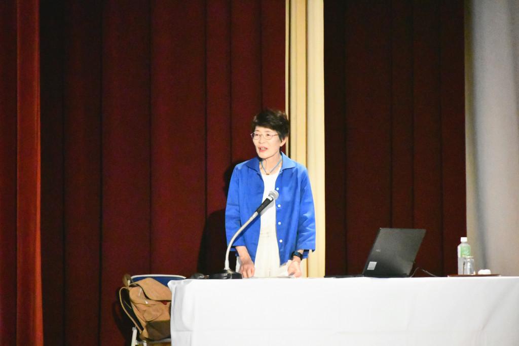 舞台上、ノートパソコンを開いた机でマイクに向かって立ちあがって自己紹介をする藤井さんの写真