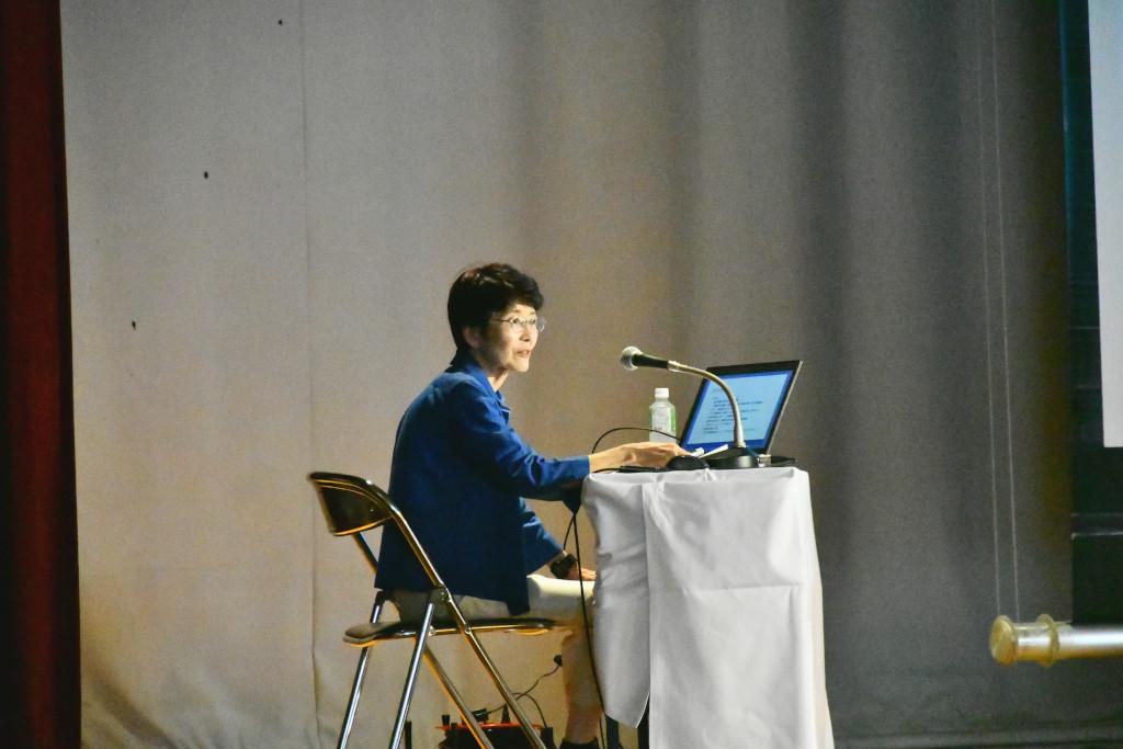 舞台上、ノートパソコンを開いた机でマイクに向かって座り、お話を進める藤井さんの写真