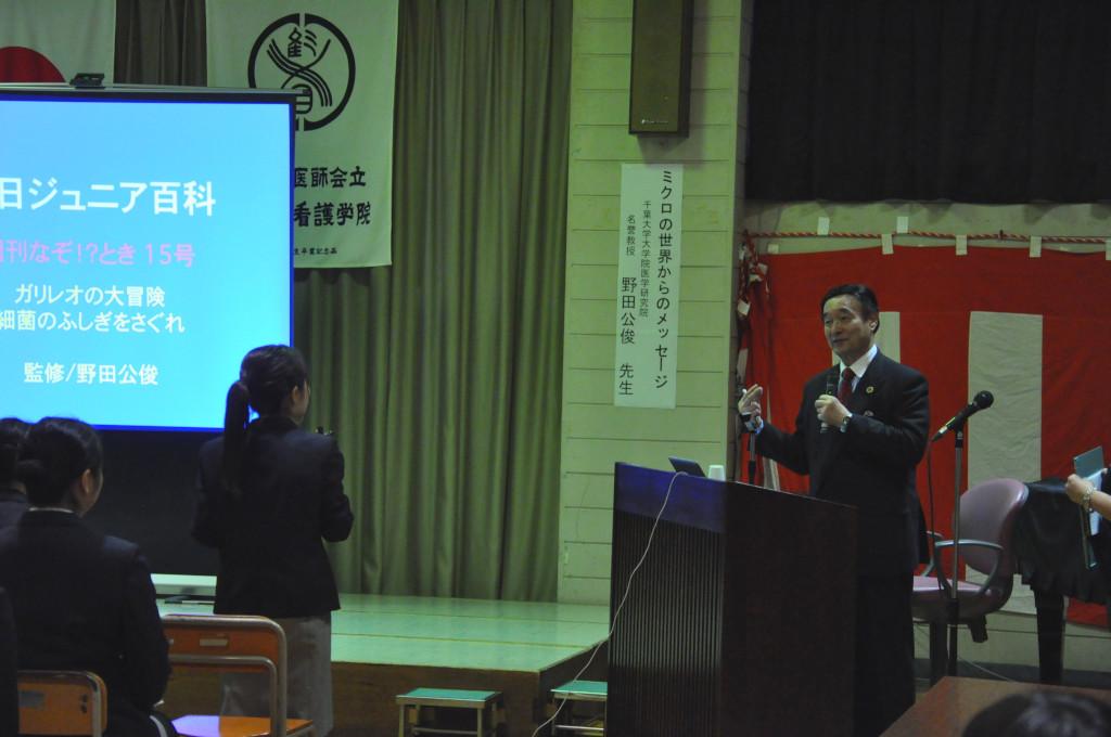 スクリーンの横に立ってマイクを持って話をしている女子生徒と野田先生の写真