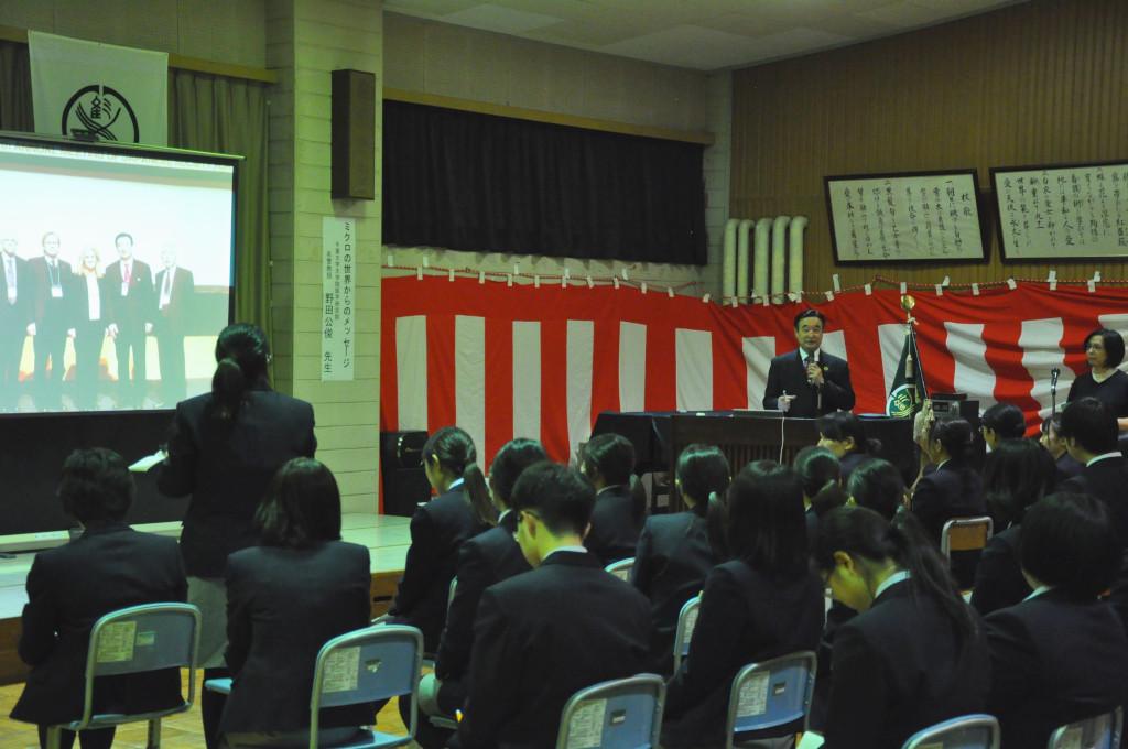 数人の人が写ったスクリーンの横に立って話をしている野田先生とマイクを持って話をしている女子生徒の後姿の写真