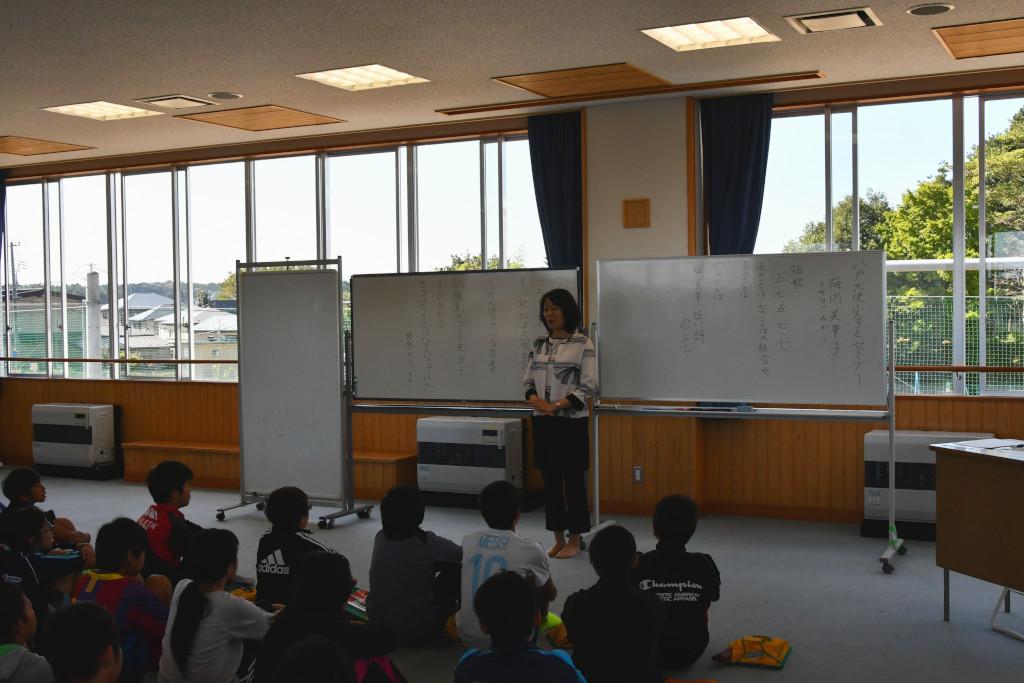 大きな窓の教室でホワイトボードを背に、床に座る児童の前で自己紹介をする梅内さんの写真