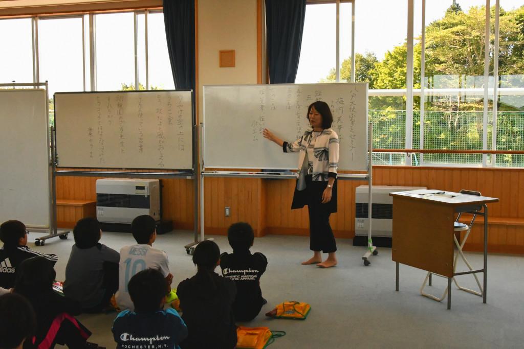 大きな窓の教室でホワイトボードを背に、床に座る児童の前でホワイトボードに手をやりながら説明をする梅内さんの写真