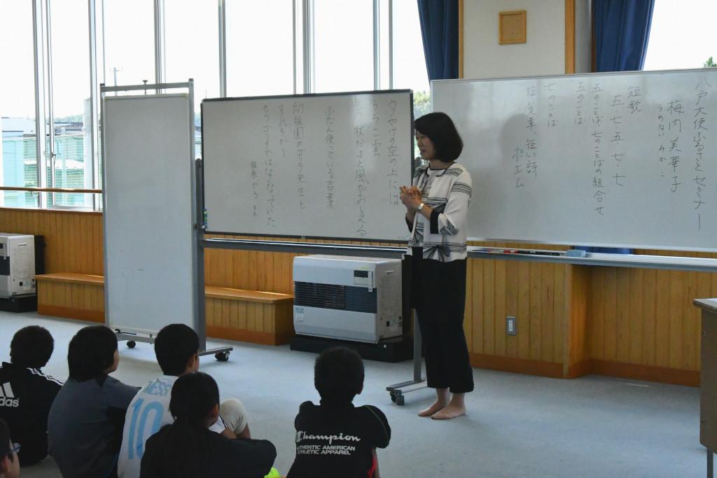 大きな窓の教室でホワイトボードを背に、床に座る児童の前でお話をする梅内さんの写真