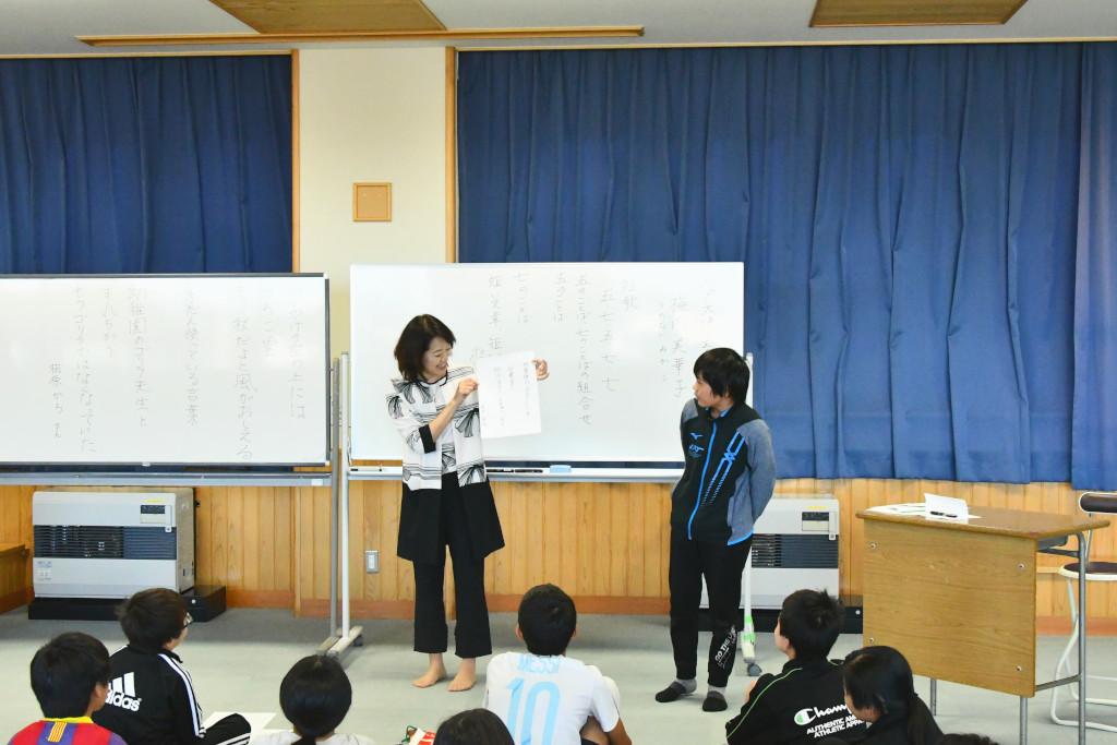 青いカーテンのひかれた教室で、生徒たちの前で男子生徒の作品の講評をする梅内さんの写真