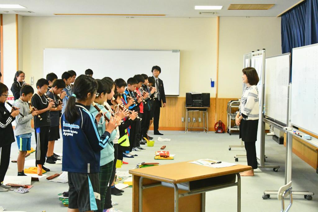 青いカーテンのひかれた教室で、ホワイトボードを背にした梅内さんにリコーダー合奏を披露する生徒たちの写真