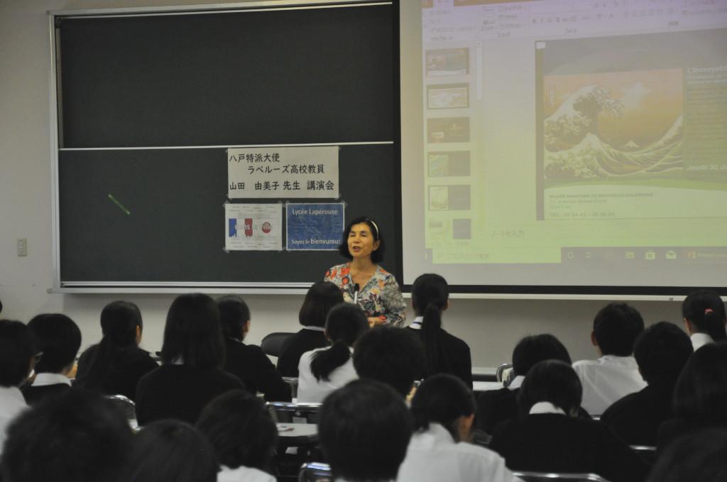 教室内、モニターの前でマイクを使い学生たちへ挨拶をする山田大使の写真