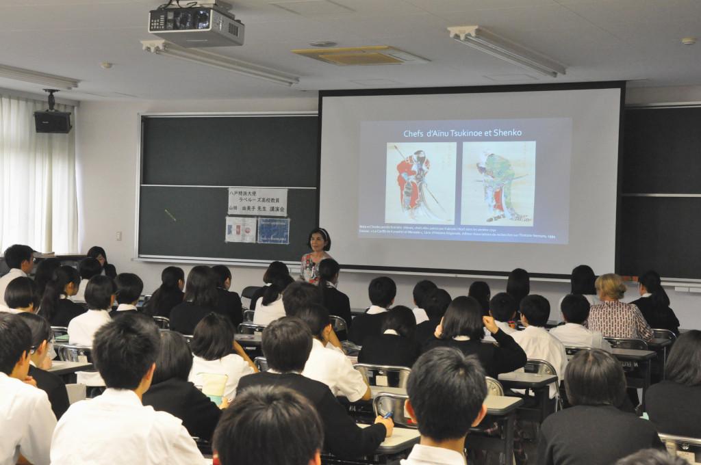 モニターに写っている2枚の画像の前で学生たちの向って話をしている山田大使の写真