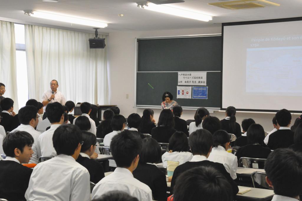 教室内、席に着いている学生たちの前でマイクを持って話している男性と、少し離れた隣に立っている山田大使の写真