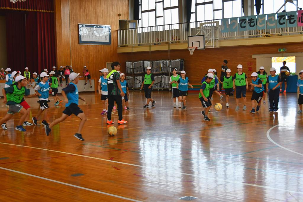 体育館にてサッカーのドリブルをしている生徒の周りでボールを追いかけようとしている生徒たちの写真